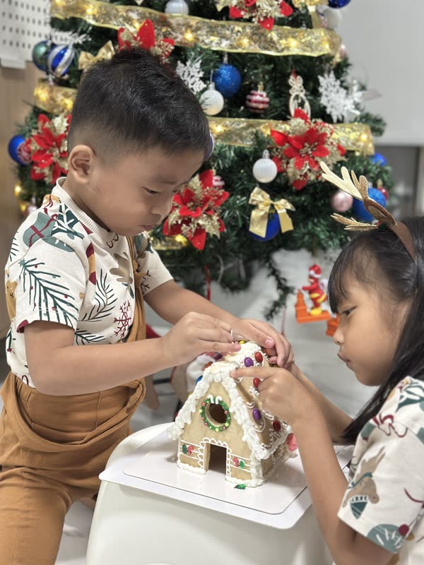 Two kids decorating their DIY cookie house together by the Christmas tree