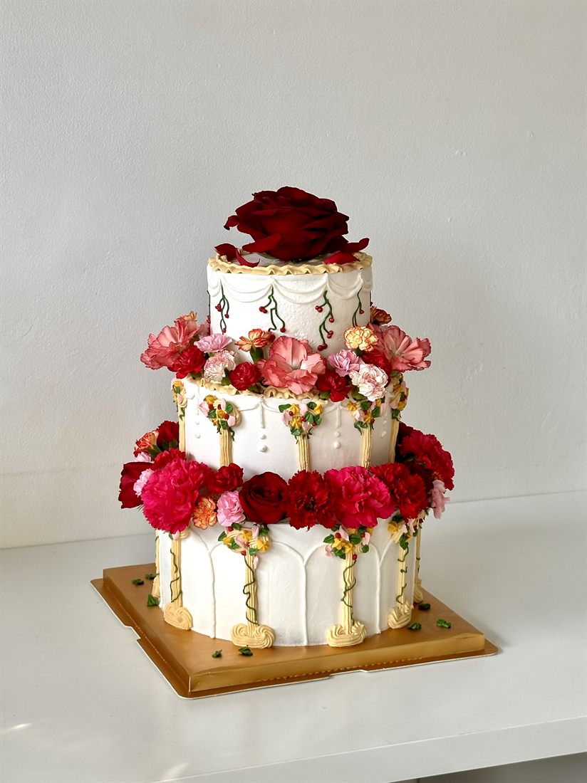 A three-tier Sugarcoat cake with hand-piped arches, gold columns and real red and pink blooms, crowned with a deep red rose