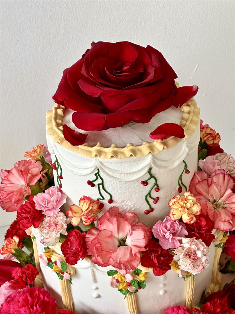 The crown of a Sugarcoat tiered cake — a deep red rose over gold-trimmed buttercream and real blooms